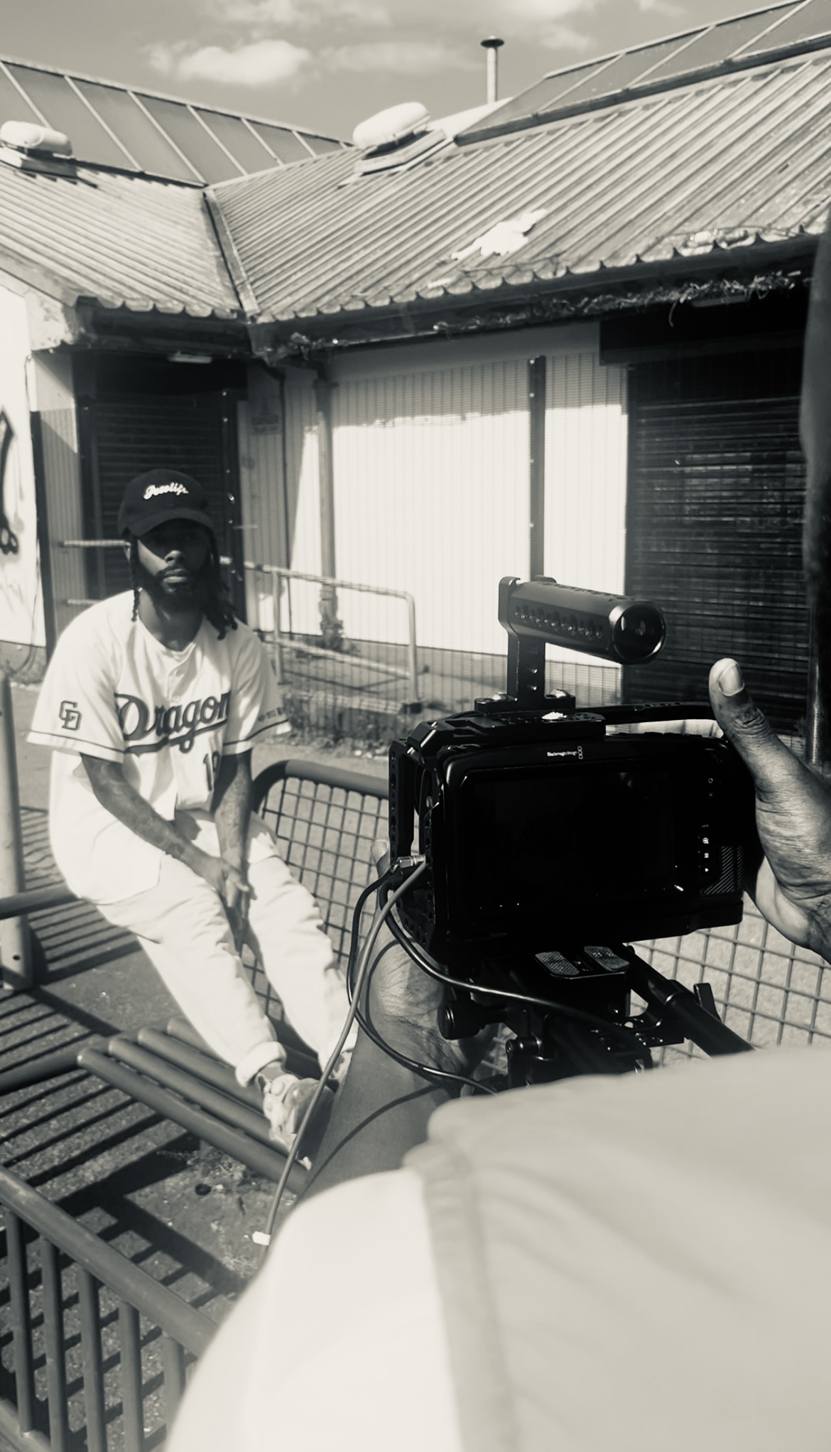 Image showing a man sitting on the table of a metal bench, a building stands behind him and he is being captured on a video camera by a man that we can see in the foreground.